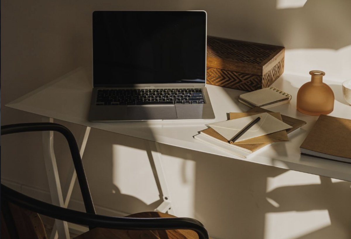Mesa de trabalho de um consultório na luz da manhã: laptop fechado de tela apagada, caderno com envelopes e uma caneta apoiada, caixa de madeira entalhada, vaso âmbar e sombras de janela sobre a mesa branca.